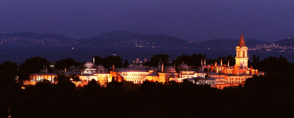Topkapi Palace illuminated at night with glowing domes and tower against a dark sky and distant Bosphorus backdrop